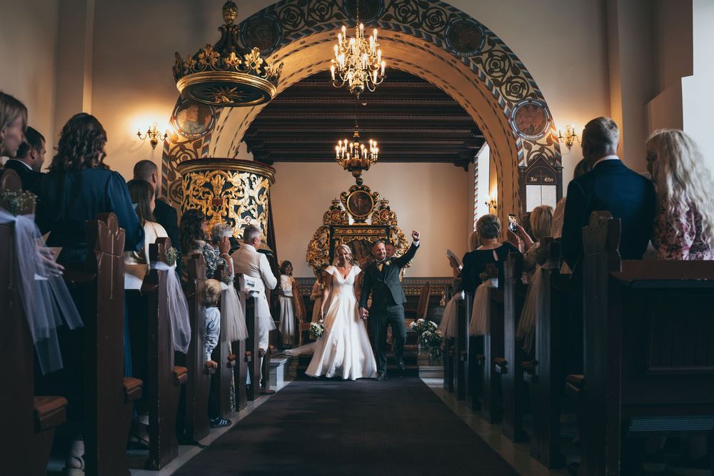 Bride and groom walk down the aisle of an ornate church surrounded by wedding guests during a ceremony.