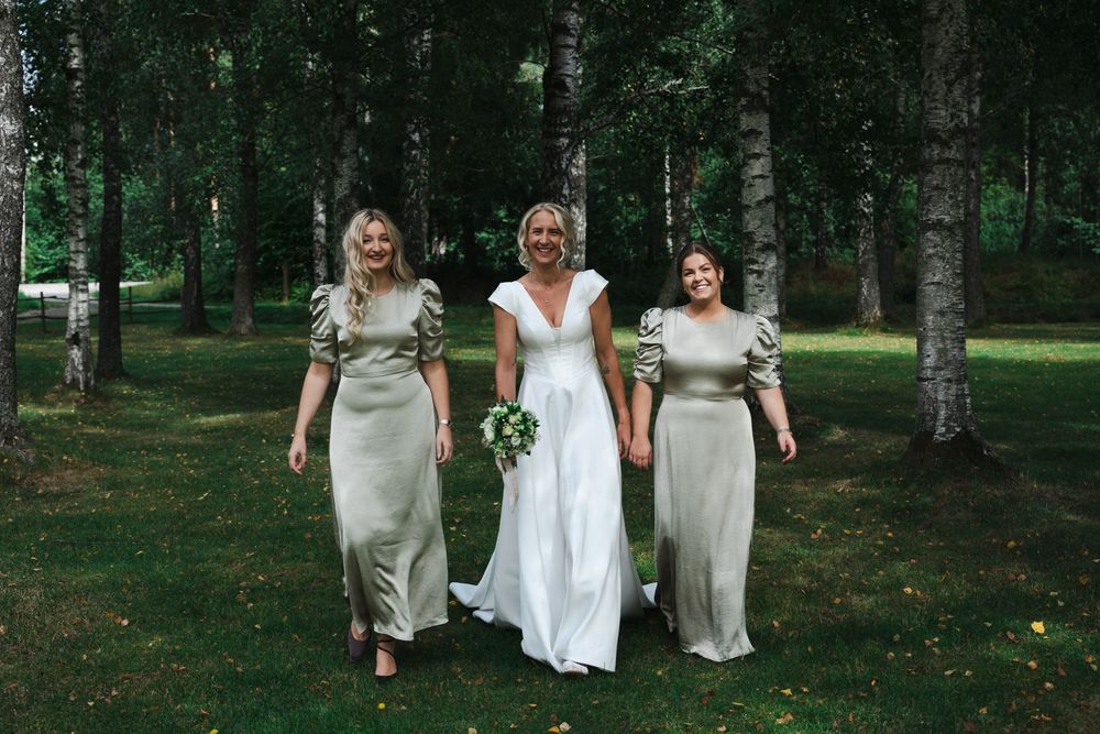 Bride in white gown walks with two bridesmaids in sage satin dresses among birch trees outdoors.