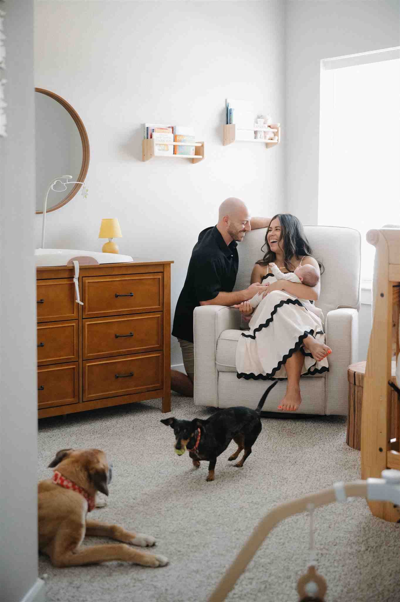A cozy nursery room with wooden furniture, white walls, and two dogs lounging on the carpet as a family relaxes in an armchair.