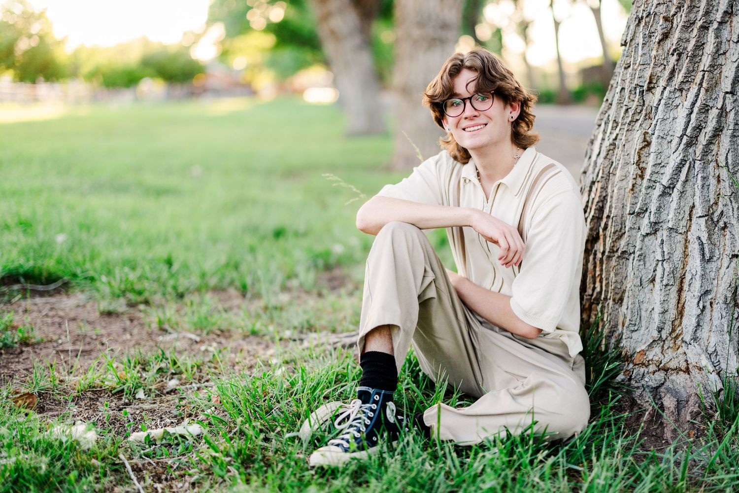 A casual portrait of someone sitting under a tree wearing neutral tones and sneakers.