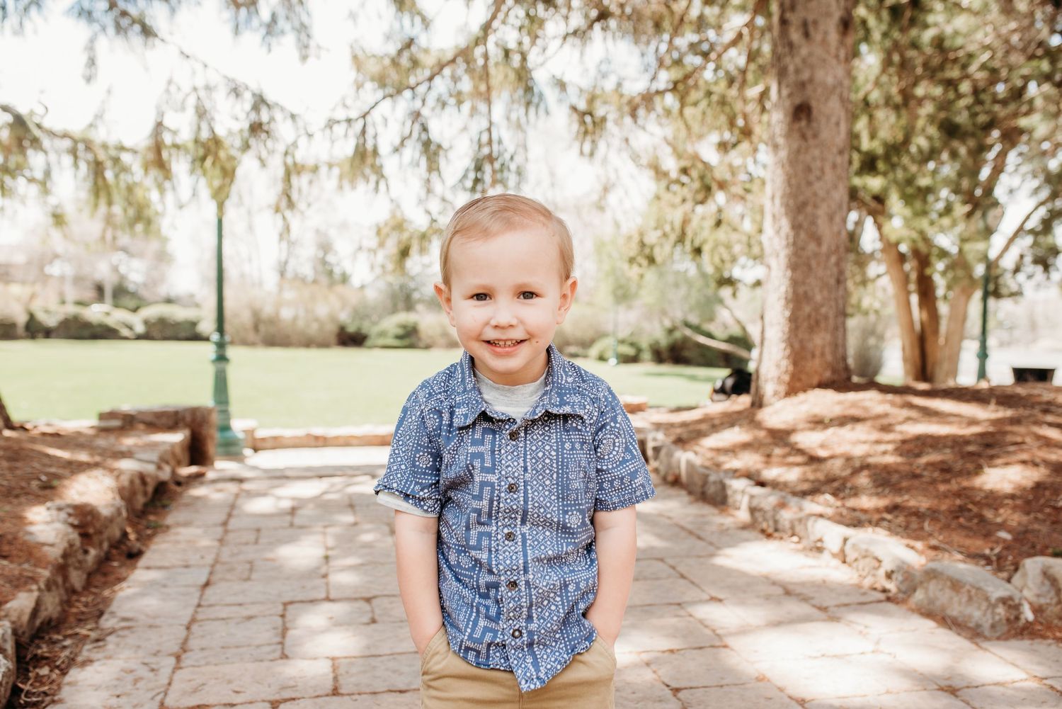 Child in denim shirt smiling on stone pathway under evergreen trees.