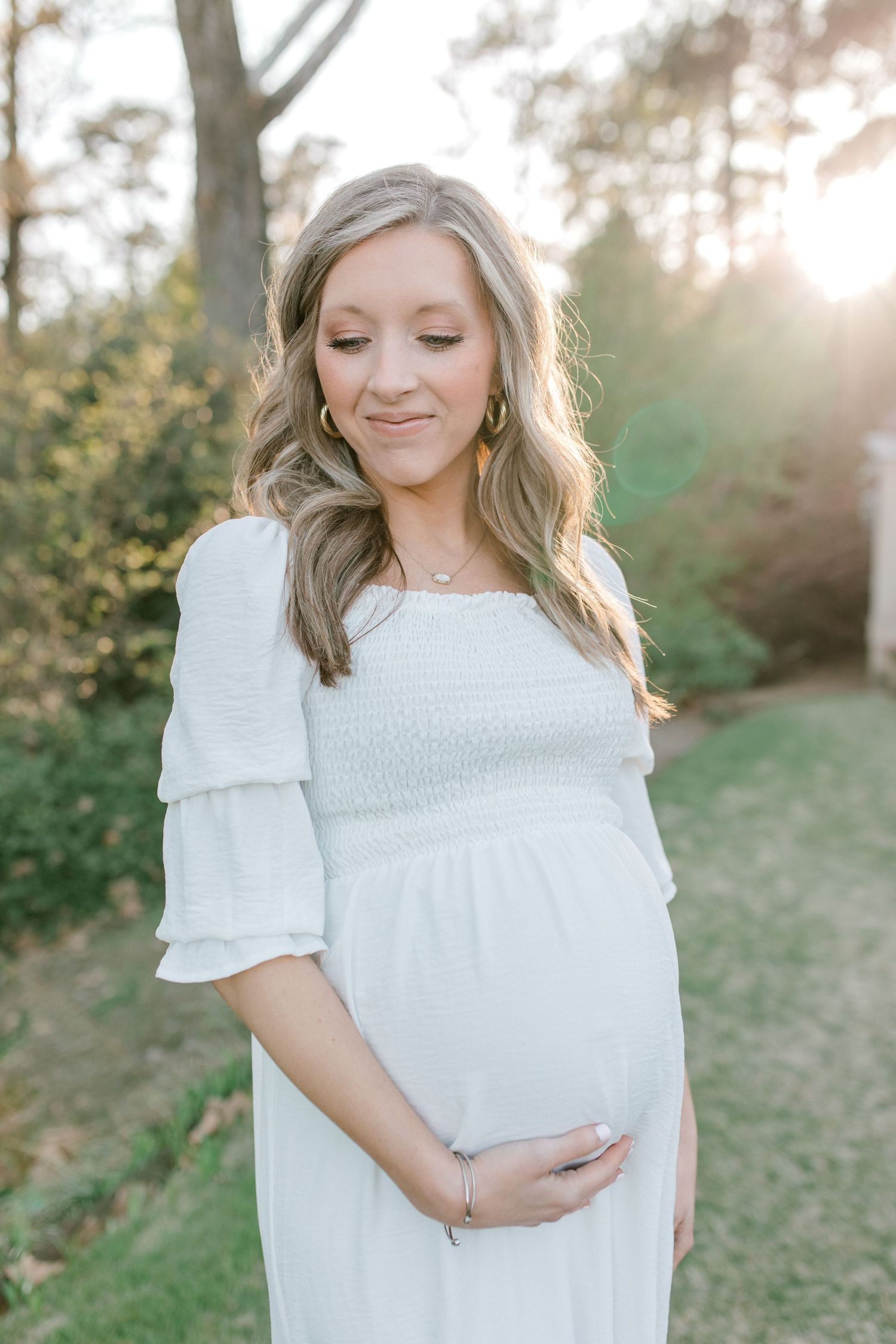 Expecting mother in white dress poses in sunlit woodland setting.