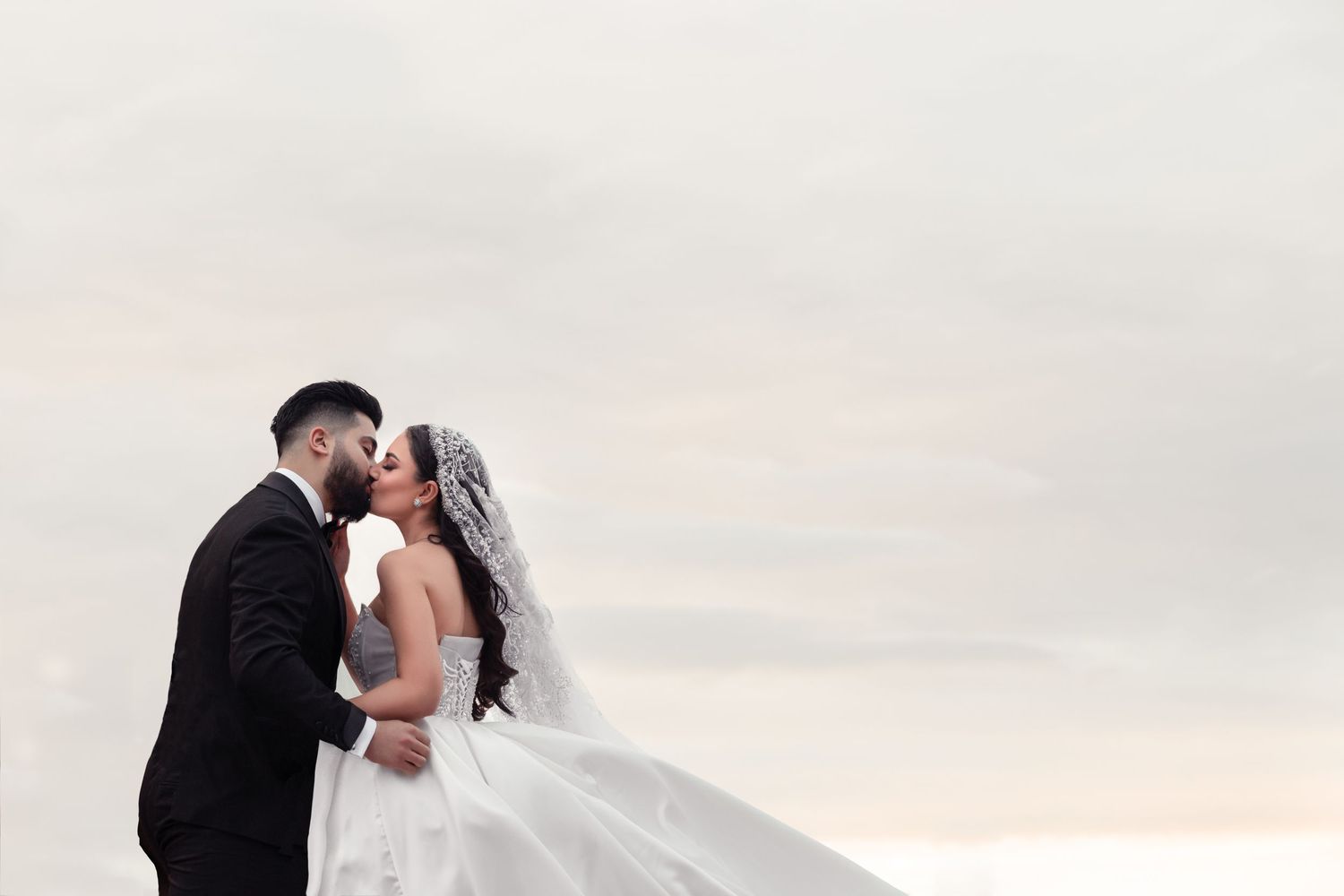Bride and groom share a romantic kiss outdoors with a flowing white veil against a soft sky.