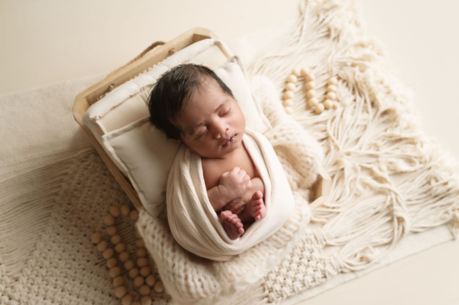 Newborn baby wrapped in cream colored blankets resting peacefully on soft white textured backdrop.