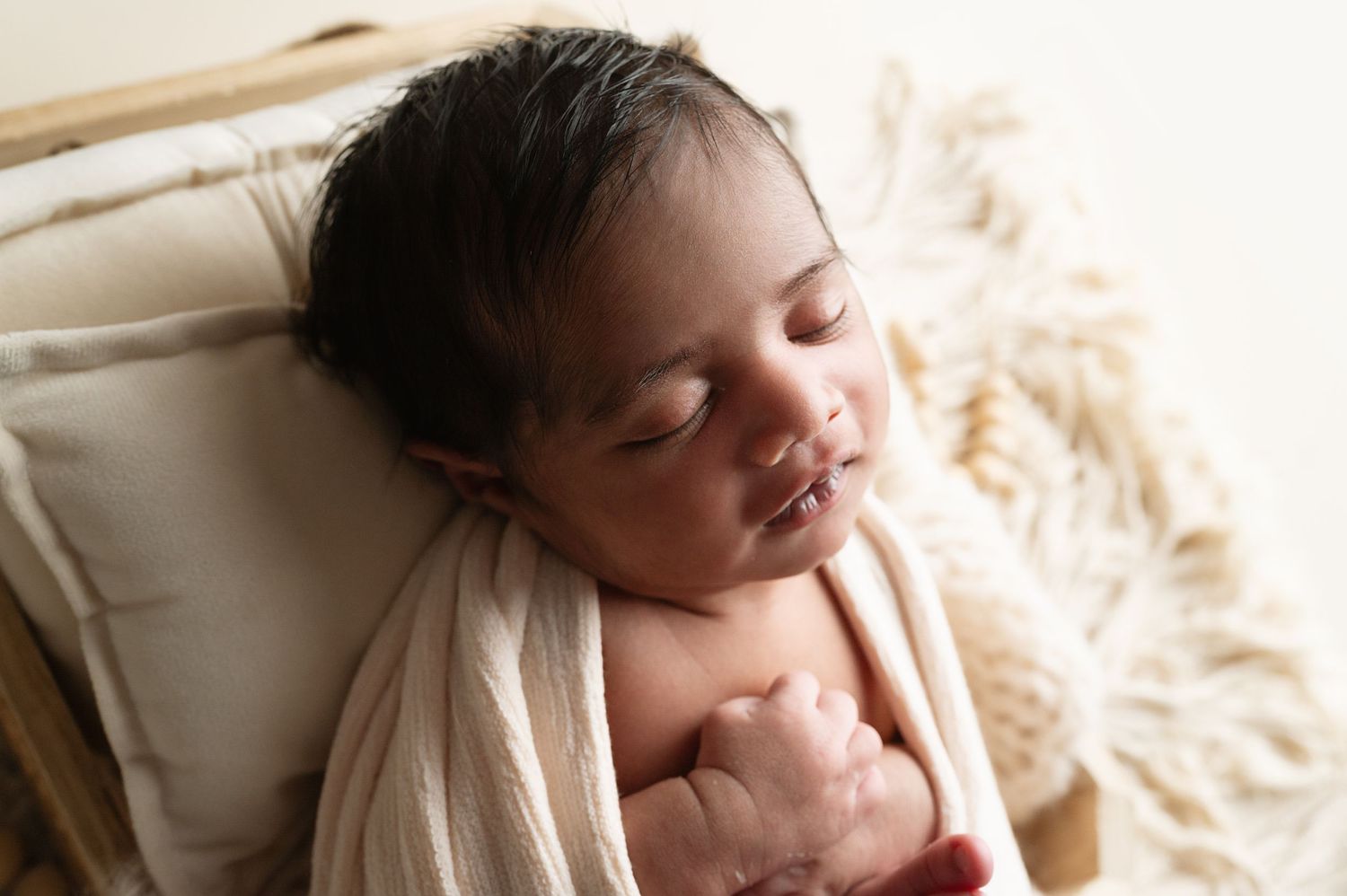 Sleeping newborn nestled in white blanket against cream colored fabric background.