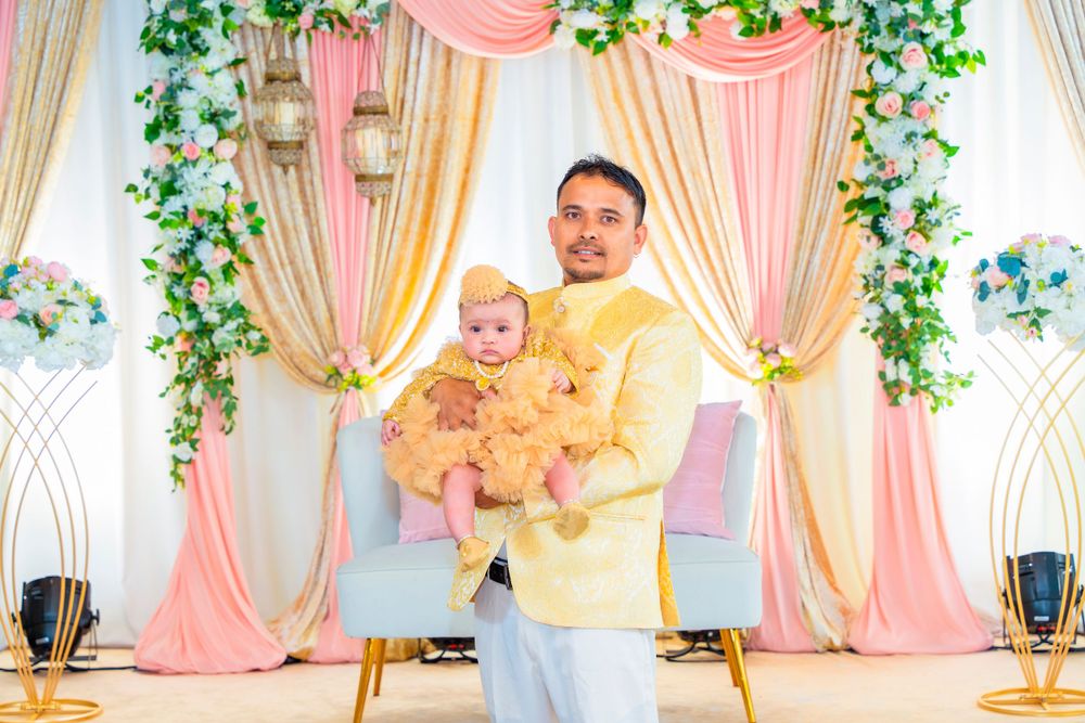 Person in yellow shirt holds baby against peach and gold draped backdrop with greenery.