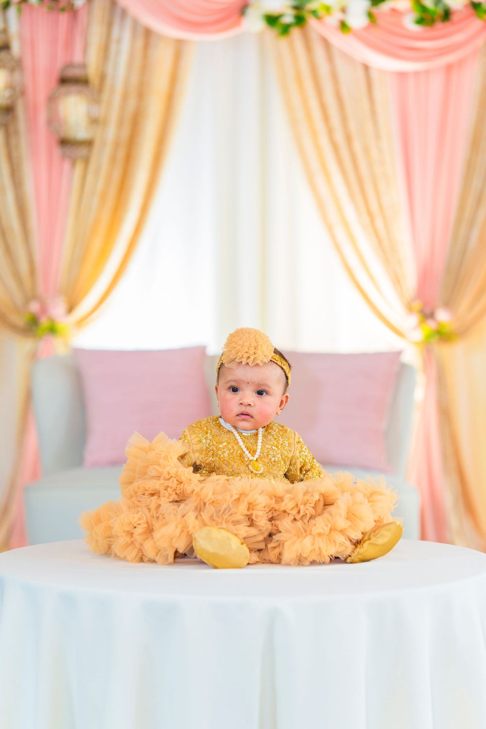 Baby in gold attire sits on white table with peach and cream draped backdrop and greenery.