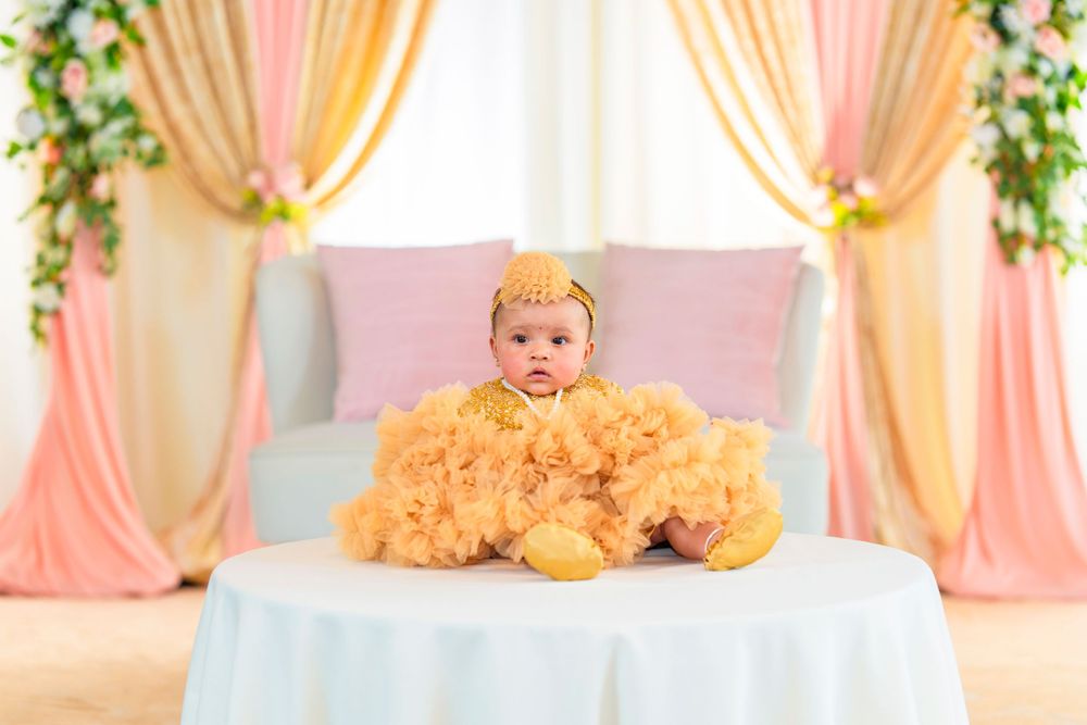 Baby wearing yellow outfit sits on white table with peach and gold draped backdrop with floral decor.