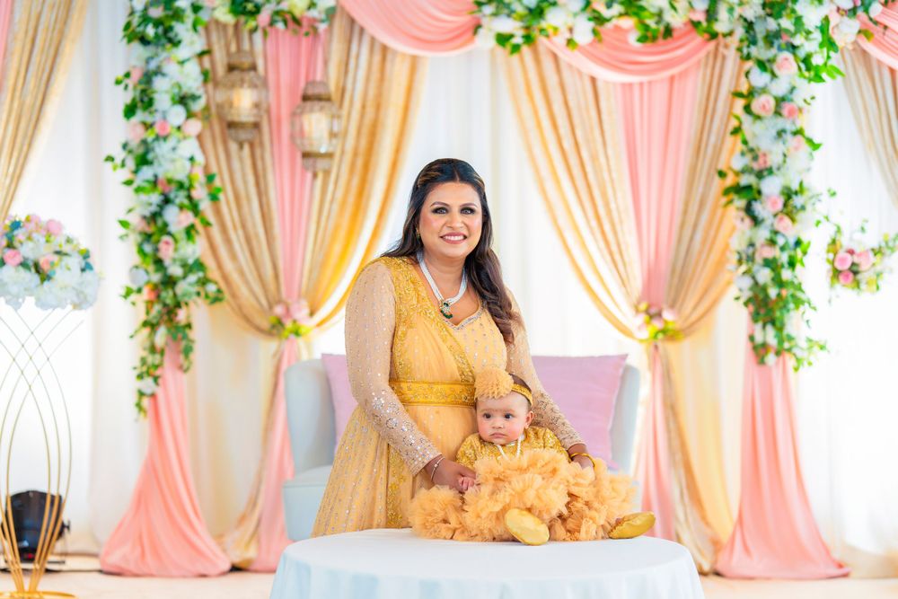 Woman in yellow dress poses with baby against peach and gold draped backdrop.