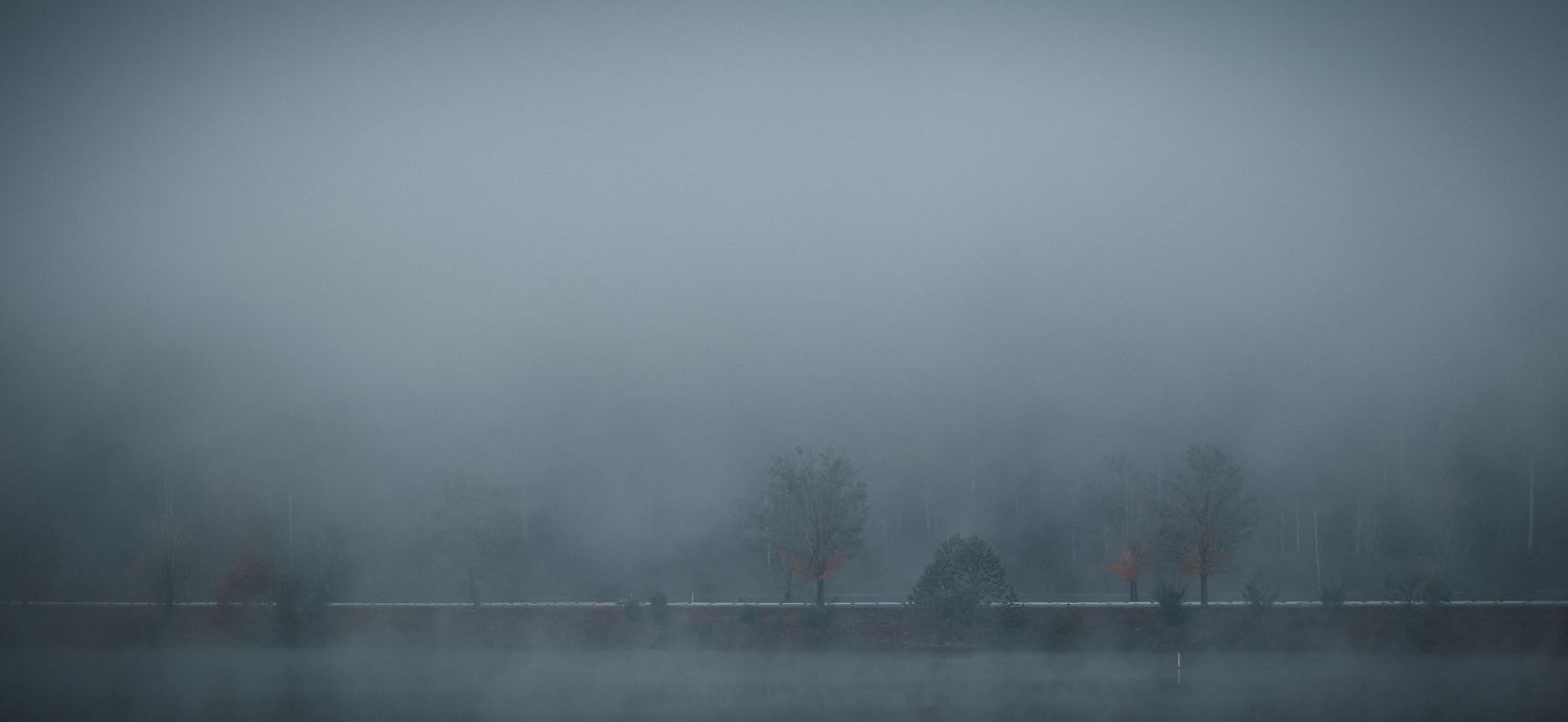 A misty lake landscape with bare trees faintly visible through thick grey fog in winter.