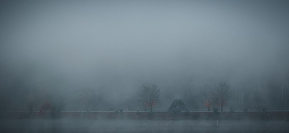 A misty lake landscape with bare trees faintly visible through thick grey fog in winter.