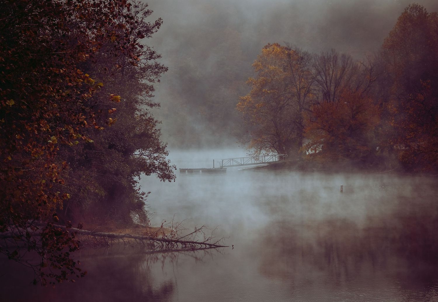 A foggy autumn river scene with mist rising over calm water surrounded by colorful fall trees.