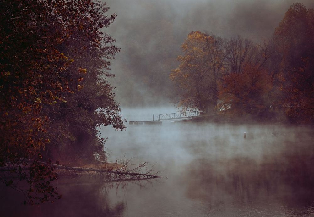 A foggy autumn river scene with mist rising over calm water surrounded by colorful fall trees.