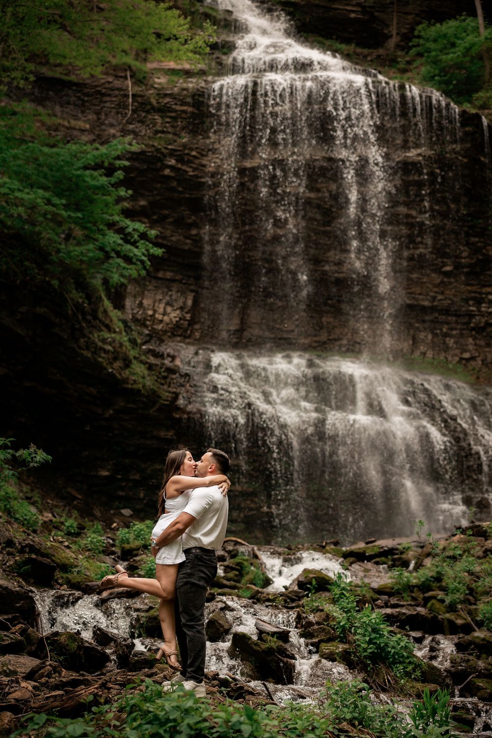 A romantic moment captured in front of a cascading waterfall surrounded by lush greenery and rocky terrain.