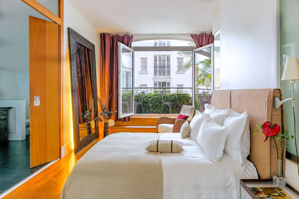 Bright bedroom with white bedding, open French doors leading to a balcony with garden views and red curtains.