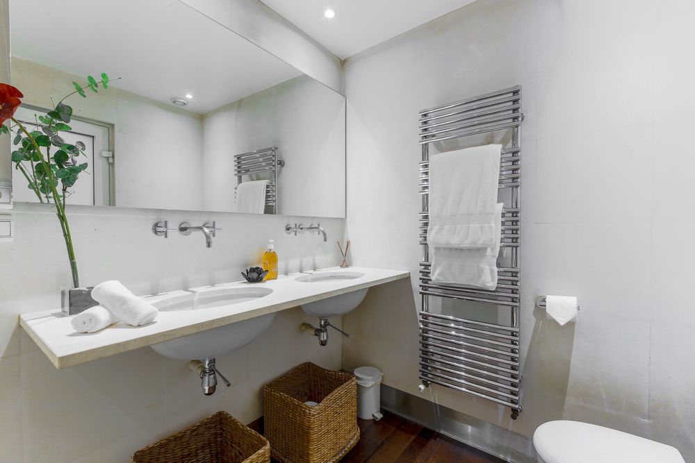 Minimalist white bathroom with wall-mounted sink, heated towel rail, wicker baskets, and fresh flower.