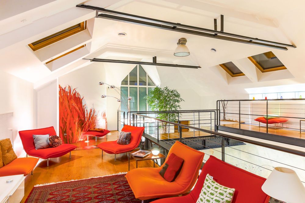 Bright attic loft lounge with skylights, vaulted ceiling, bold red and orange modern furniture, and mezzanine railing.