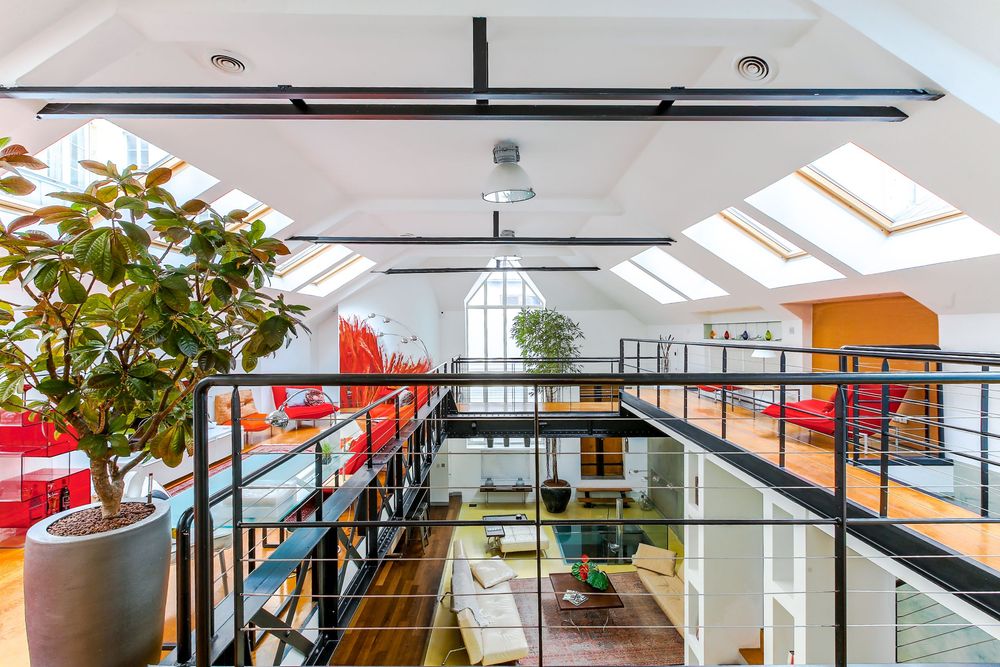 Spacious loft interior with industrial mezzanine walkway, skylights, large plant, and colorful living area below.