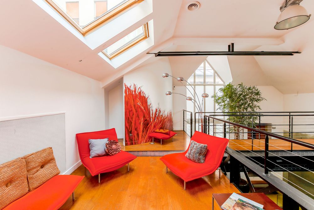 Contemporary loft lounge with red chairs, skylights, large decorative plant, and mezzanine railing.