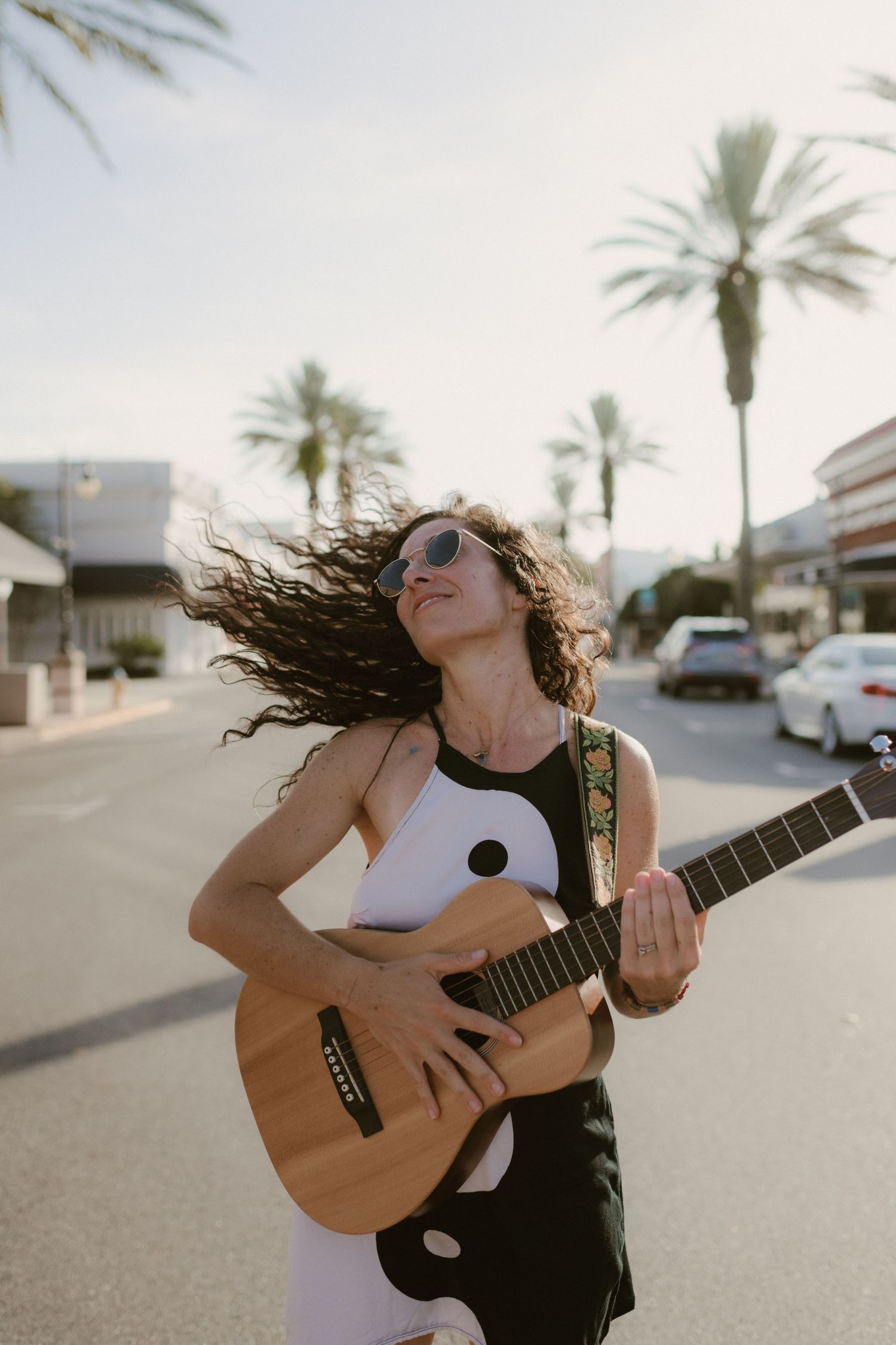 Person playing guitar on a palm-lined street with hair flowing in the wind.