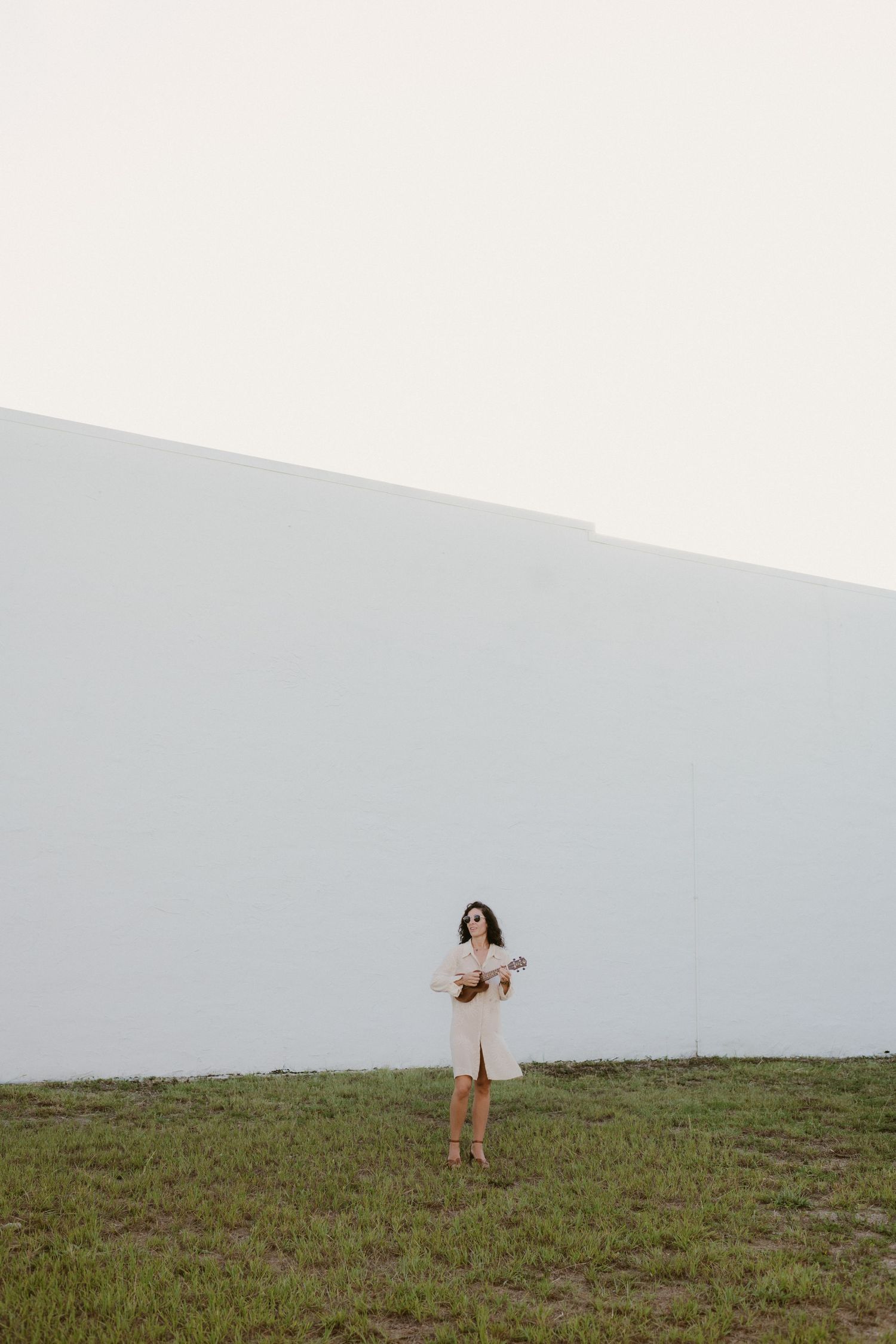 Person in white dress holding ukulele against large white wall in minimal landscape setting.
