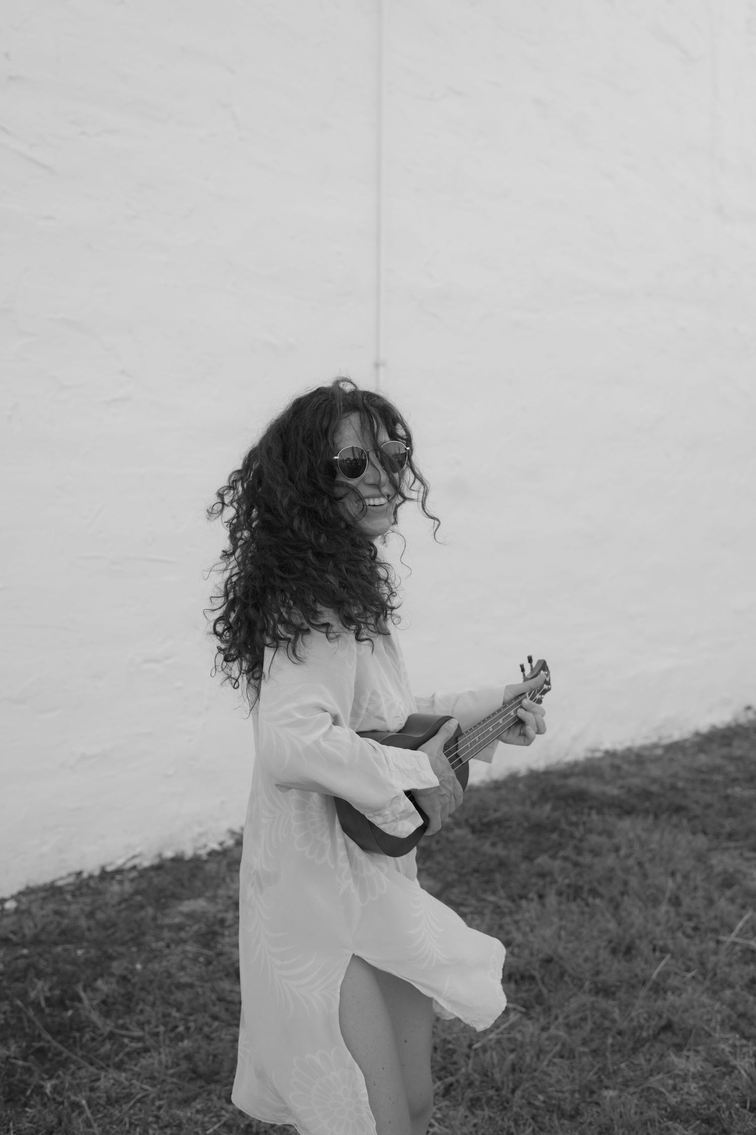Black and white image of person in flowy dress playing ukulele against white wall.