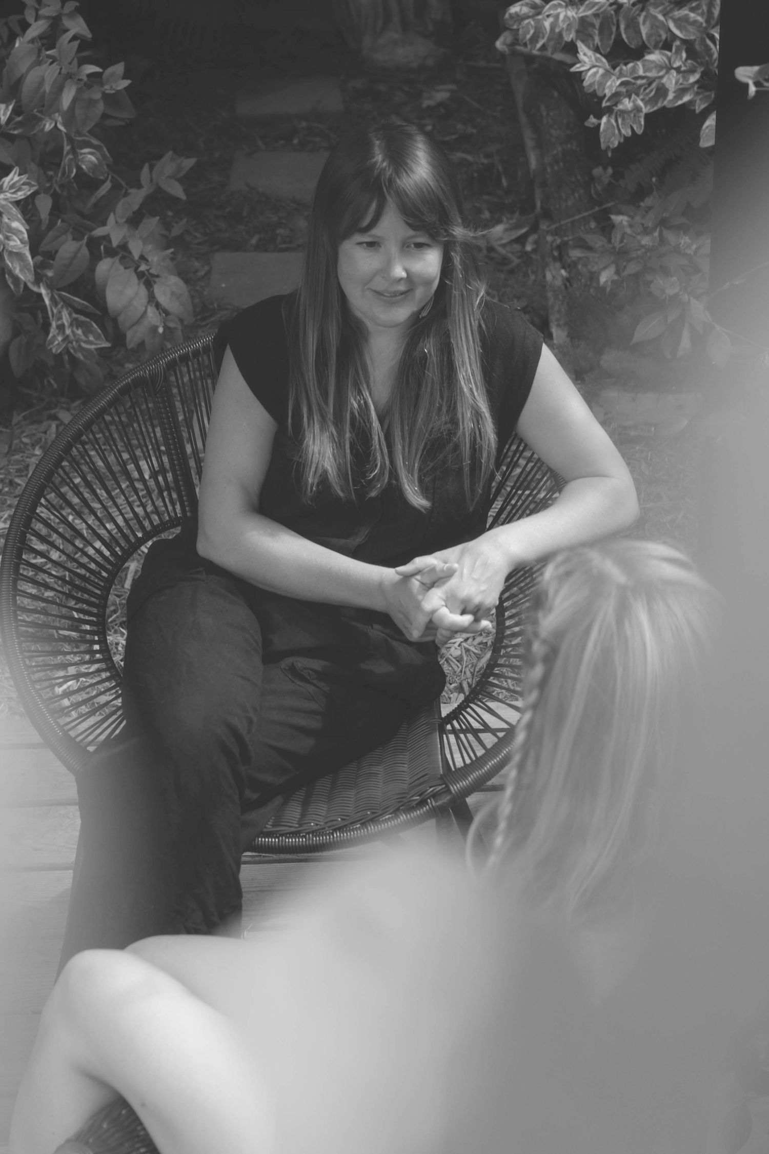 Black and white portrait of someone seated in a wicker chair surrounded by plants.