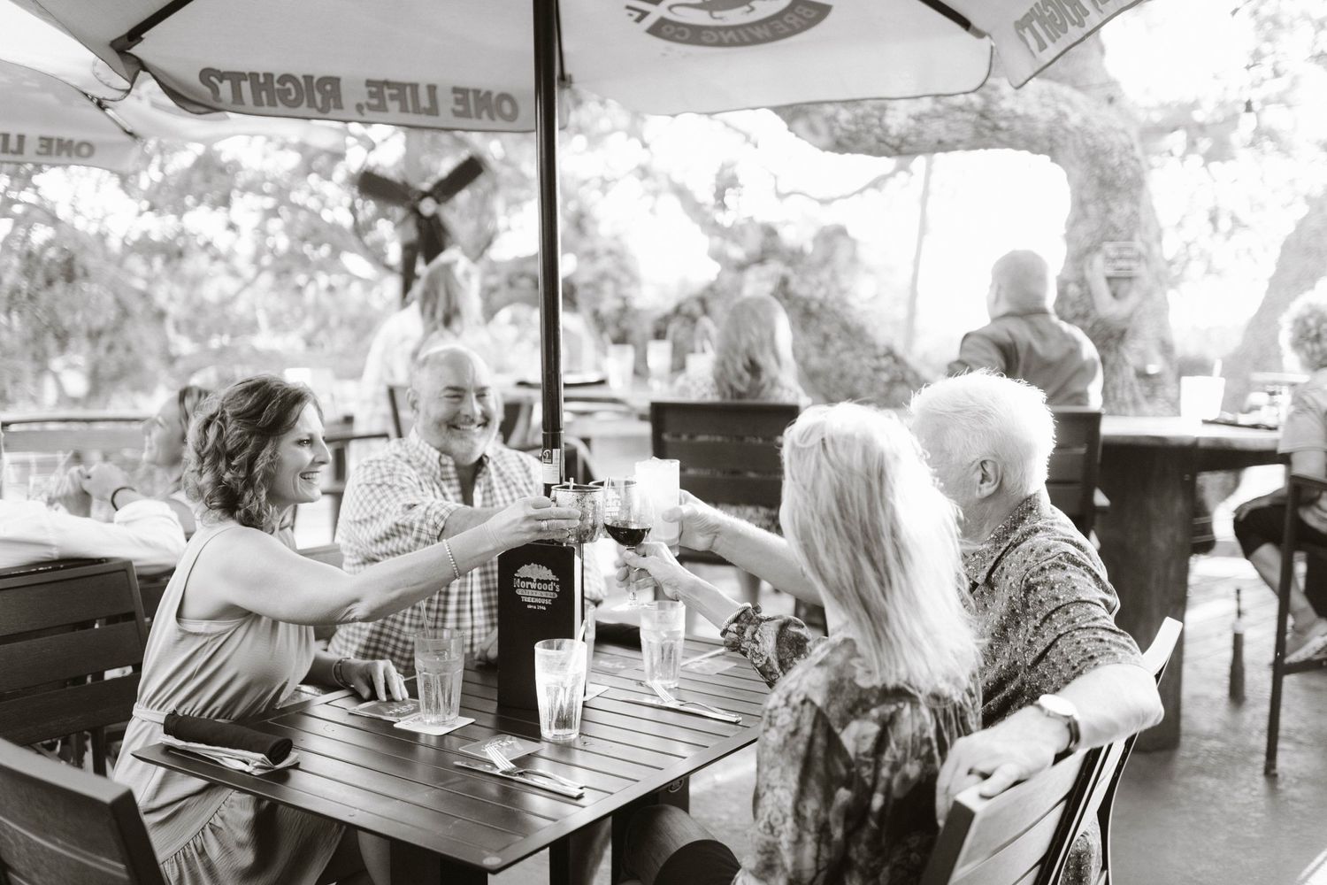 People toasting drinks at an outdoor cafe under a patio umbrella.