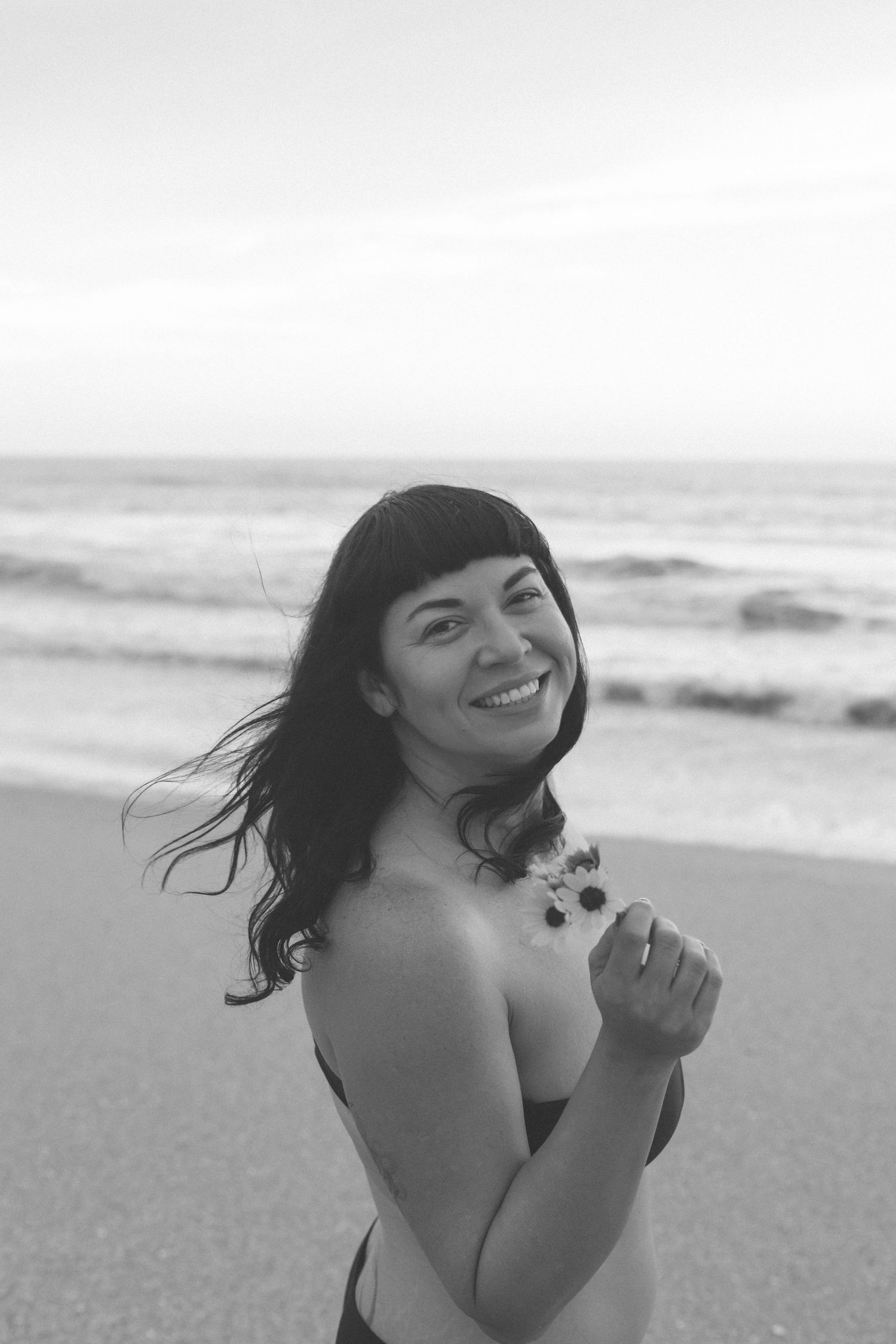 Black and white portrait of a person smiling joyfully on a beach with wind-blown hair.
