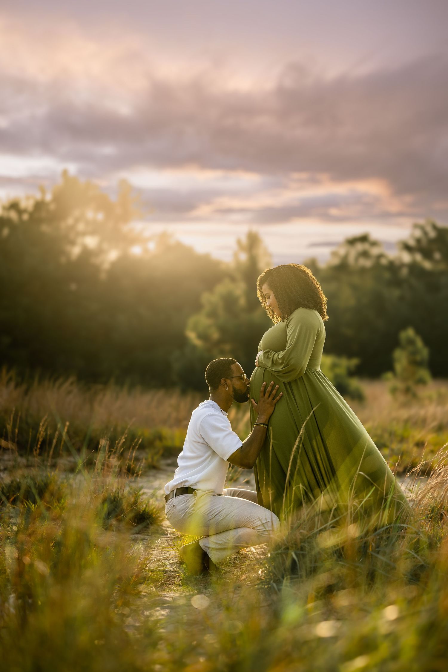 A family shares a tender moment together in a golden wheat field at sunset, wearing coordinated white and cream outfits.
