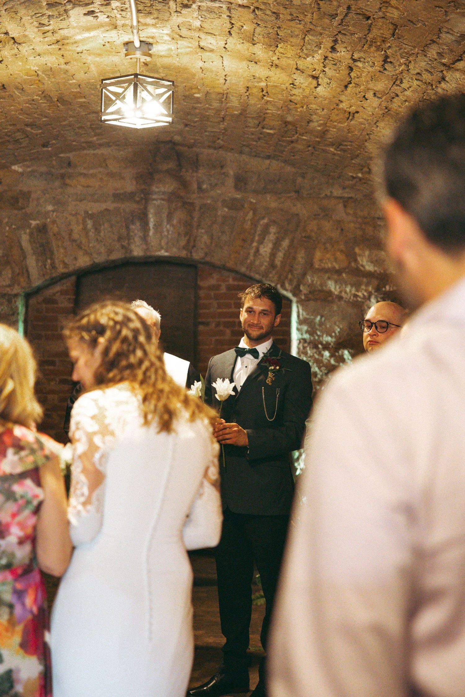 Wedding ceremony takes place in rustic brick cellar with arched ceiling and vintage lighting.
