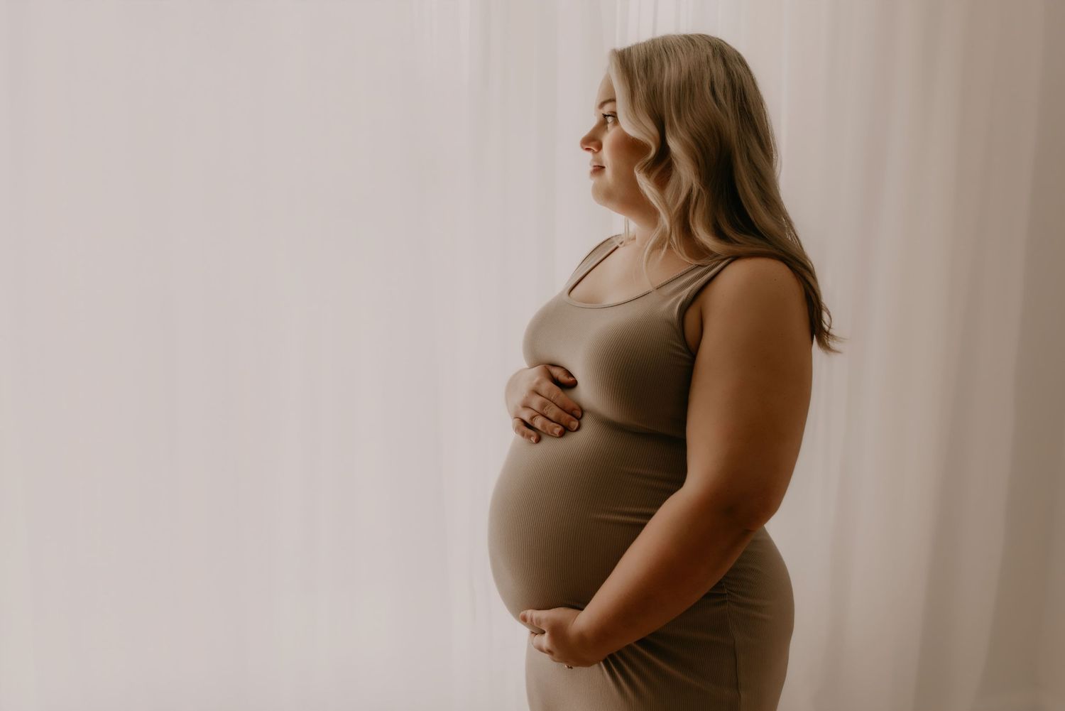 Pregnant woman in olive dress cradling her baby bump, standing by sheer white curtains in soft natural light.