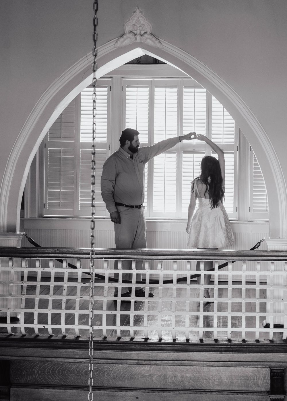 A couple poses in a church balcony with an elegant arched window frame behind them.