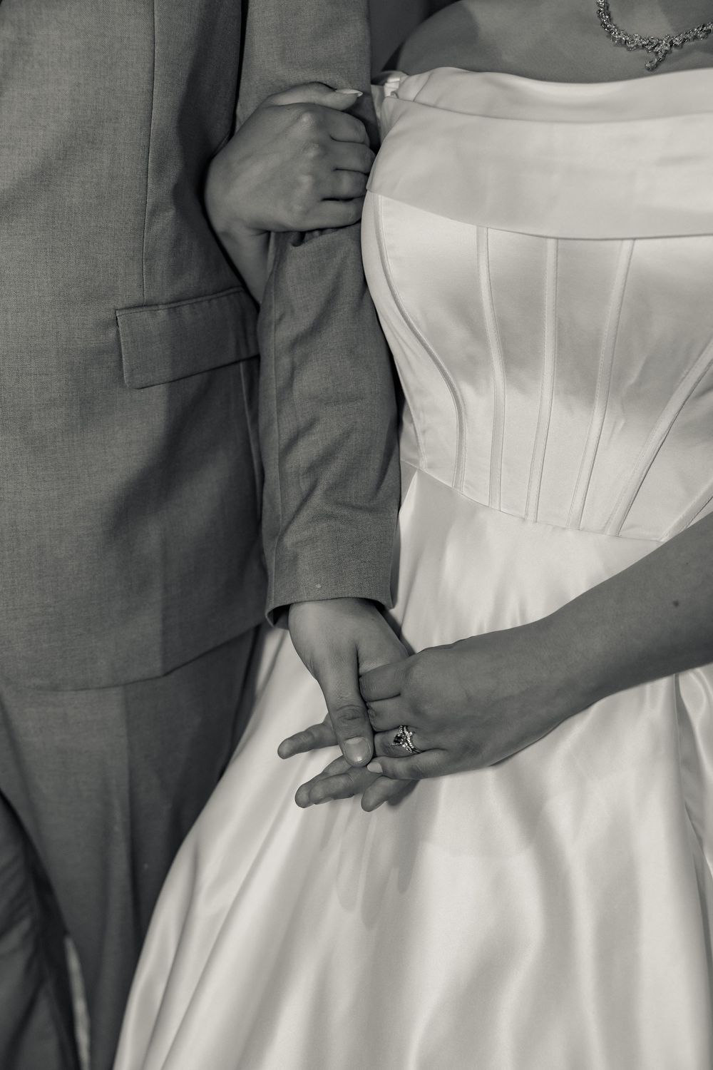 Close-up detail of hands resting on a white wedding dress against a gray suit background.