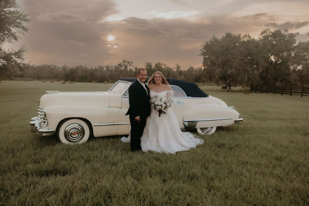Vintage white Cadillac parked in a grassy field during sunset with a couple in formal wedding attire.
