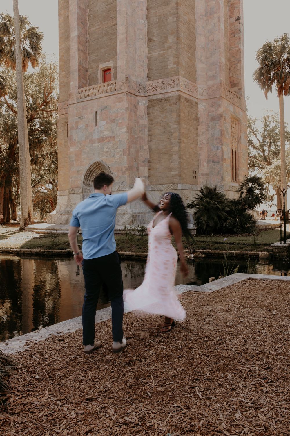 Couple dancing near a historic stone tower by a pond, woman in pink dress being twirled by man in blue shirt.