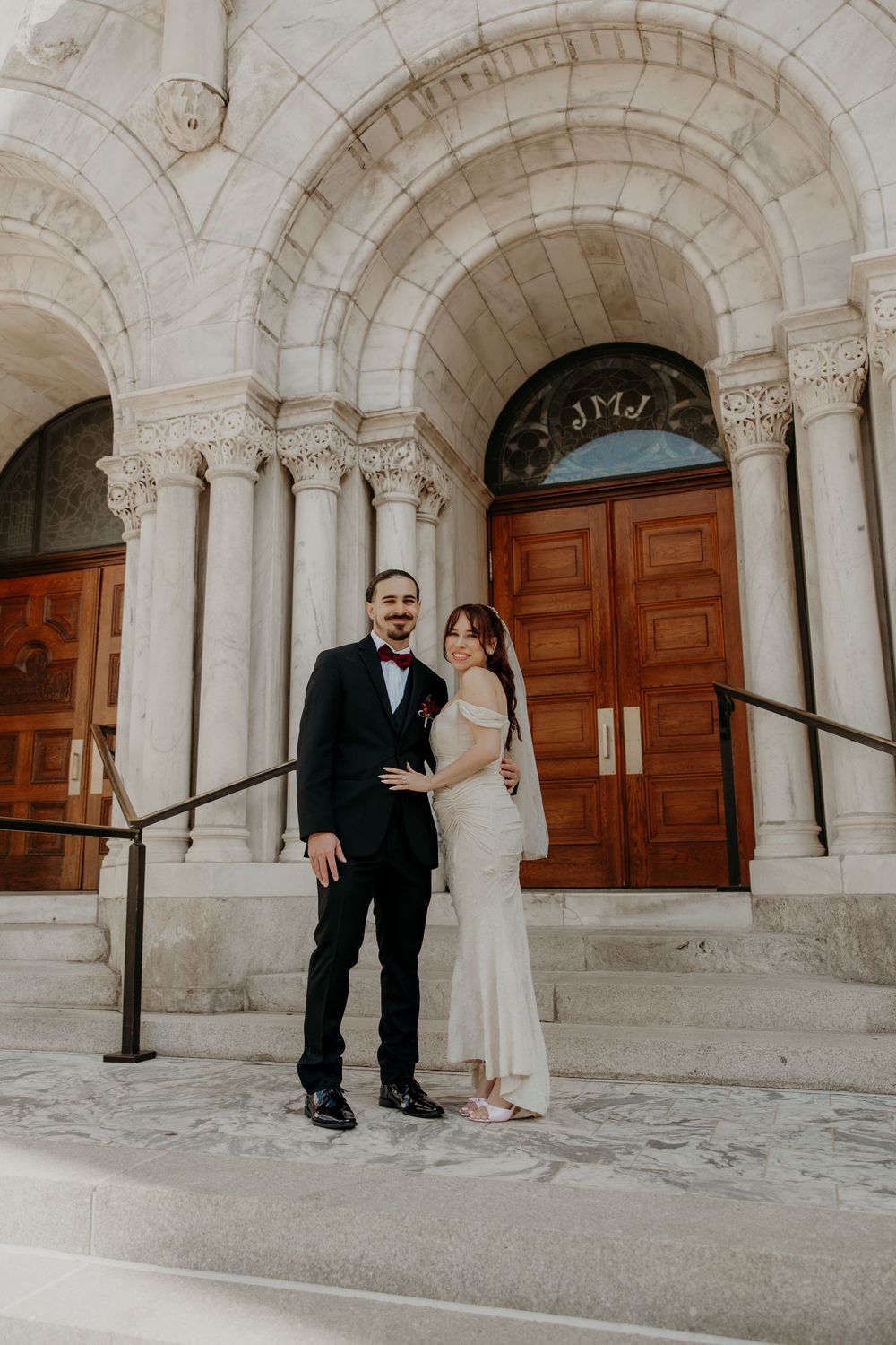 Bride and groom posing on stone steps outside a grand church with ornate arched doorway and wooden doors.