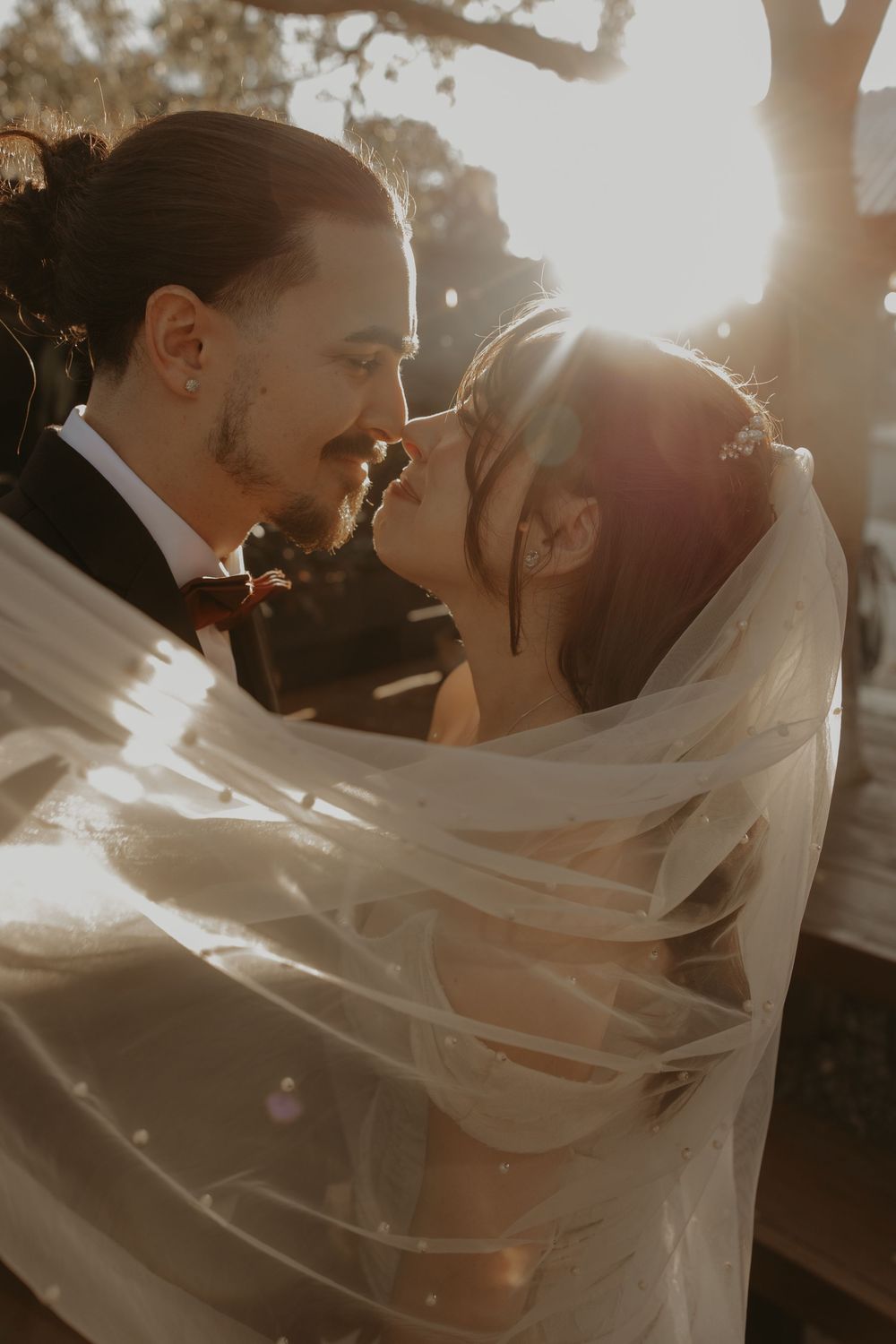 A bride and groom share a romantic moment wrapped in a flowing veil with golden sunset backlight.
