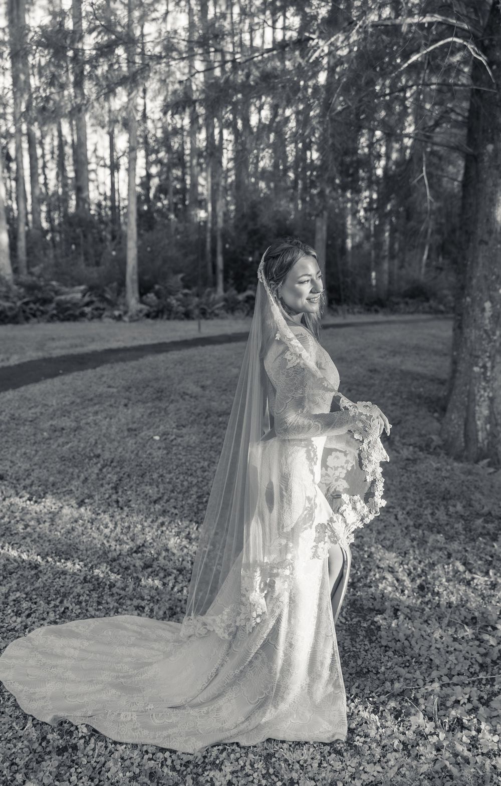 A bride in a vintage-style lace wedding gown and cathedral veil poses elegantly in a wooded outdoor setting.