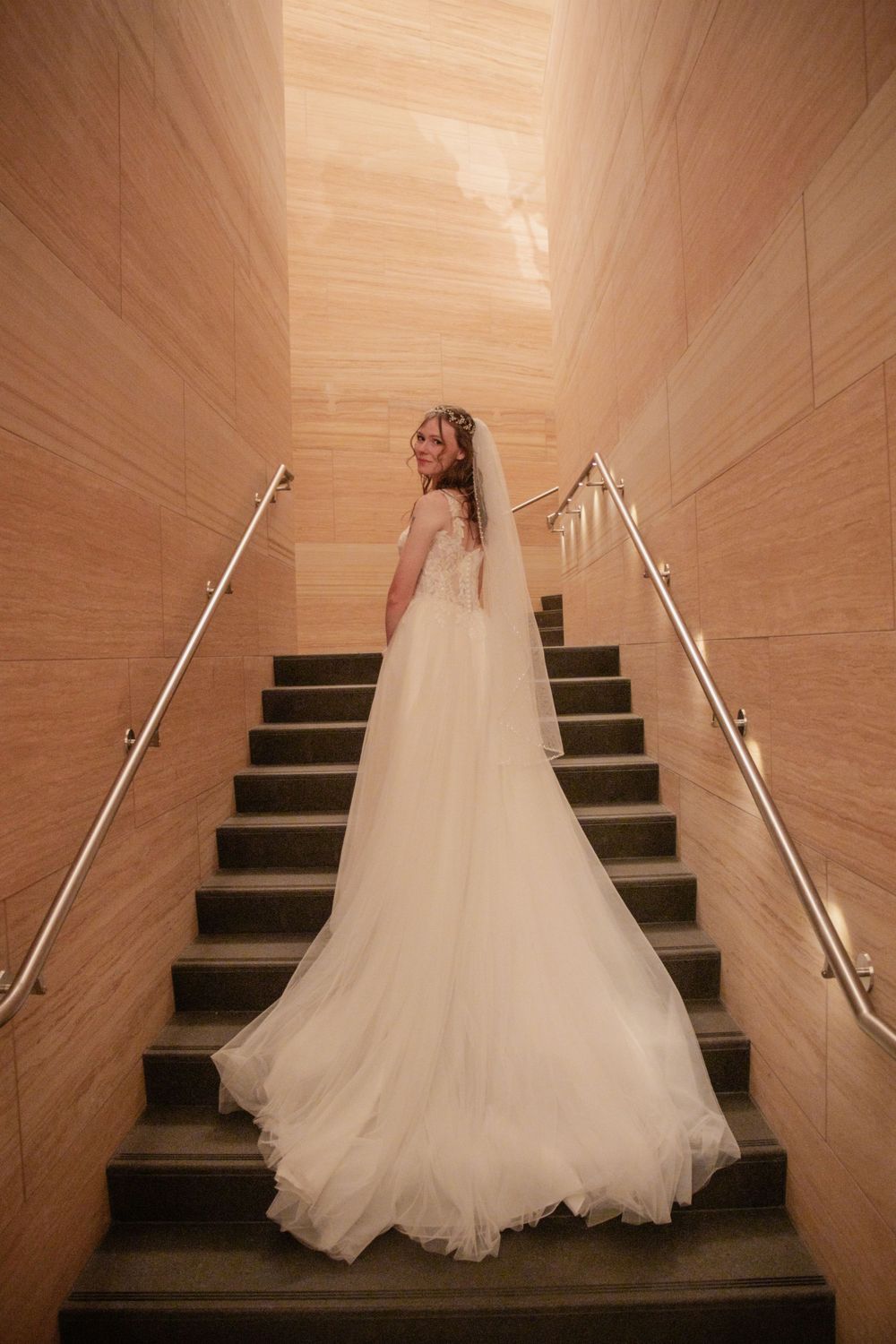 Bride in flowing white gown poses on wooden staircase with dramatic lighting.