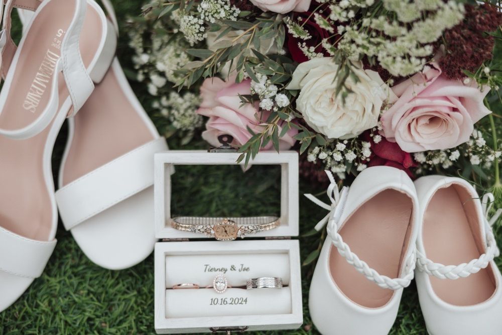 Wedding detail shot of white bridal shoes and floral ring box on grass.