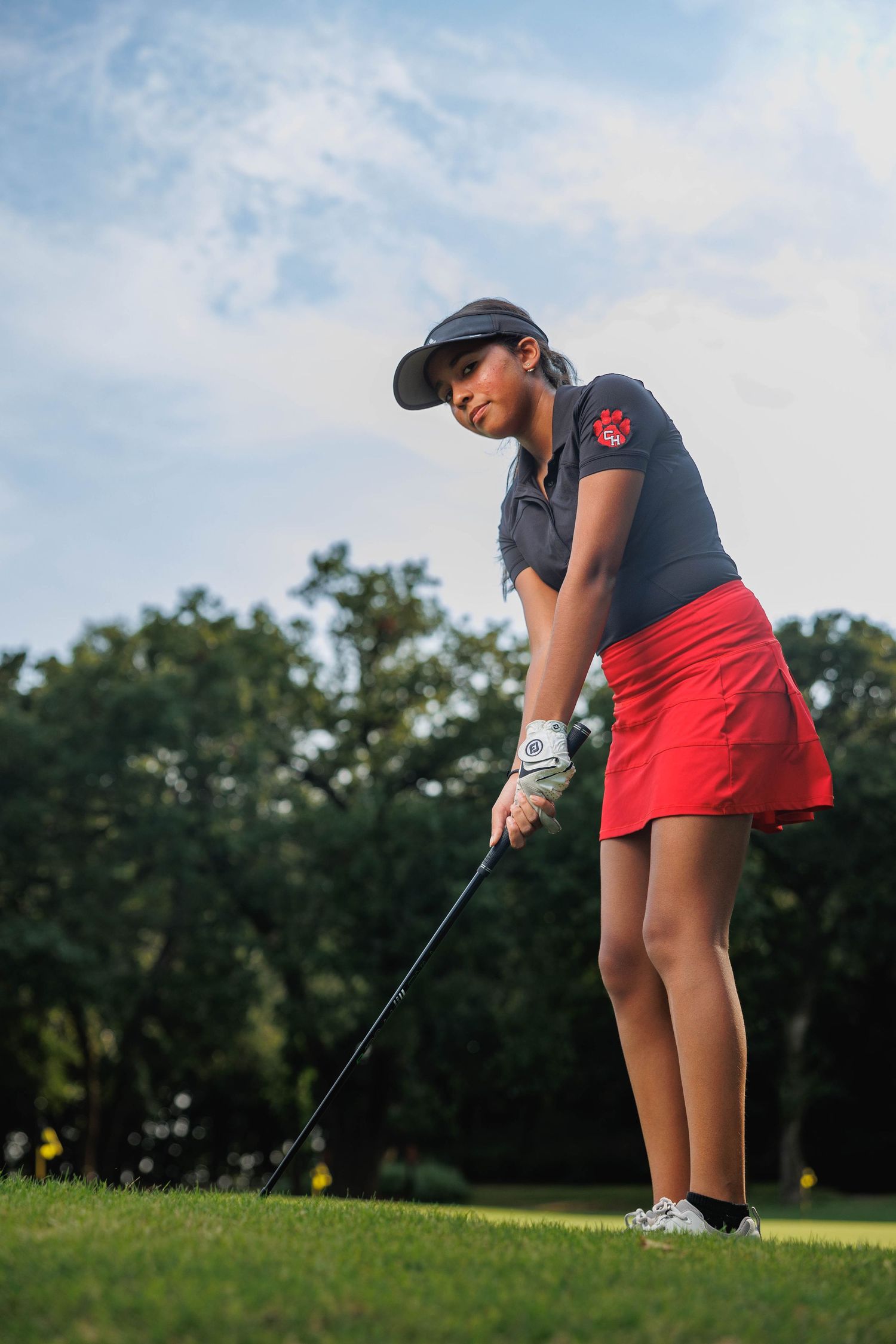 Female high school golfer in red skirt, black shirt and black visor hitting a ball out of the sand in Grapevine, TX