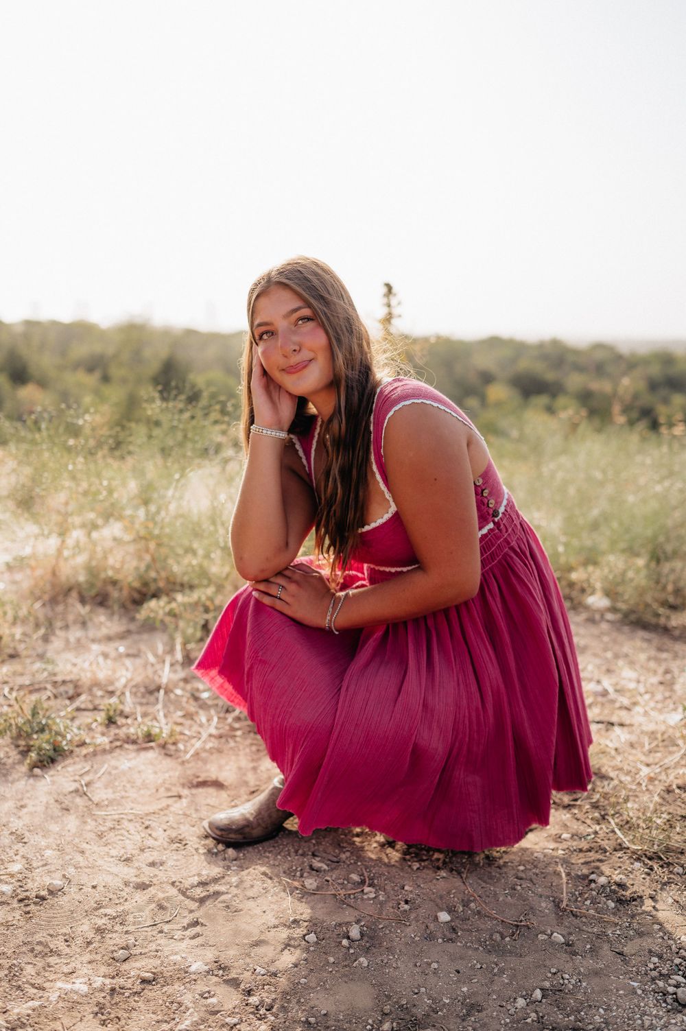 A woman in a magenta dress crouches thoughtfully on a desert path.