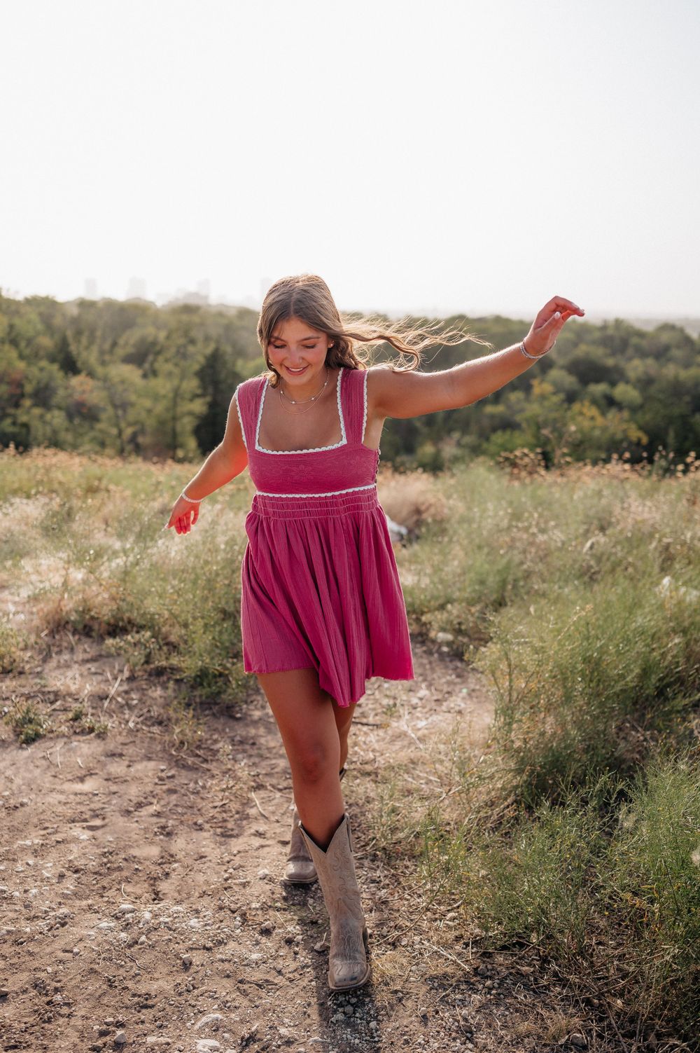 A carefree woman in a magenta dress and boots dances on a dirt trail surrounded by nature.
