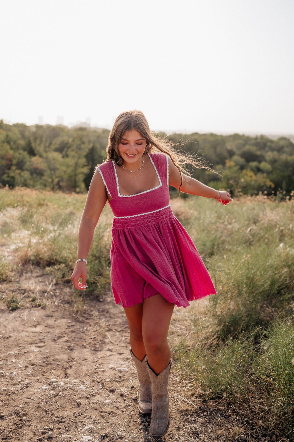 A woman twirls playfully in a flowing pink dress and cowboy boots on a hiking trail.