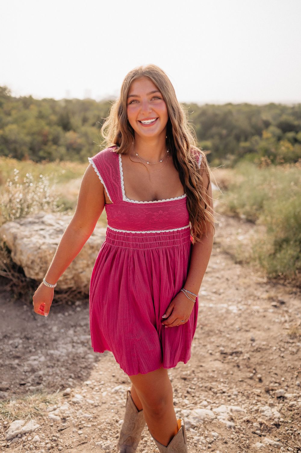 A woman in a vibrant pink dress smiles while walking along a natural trail.