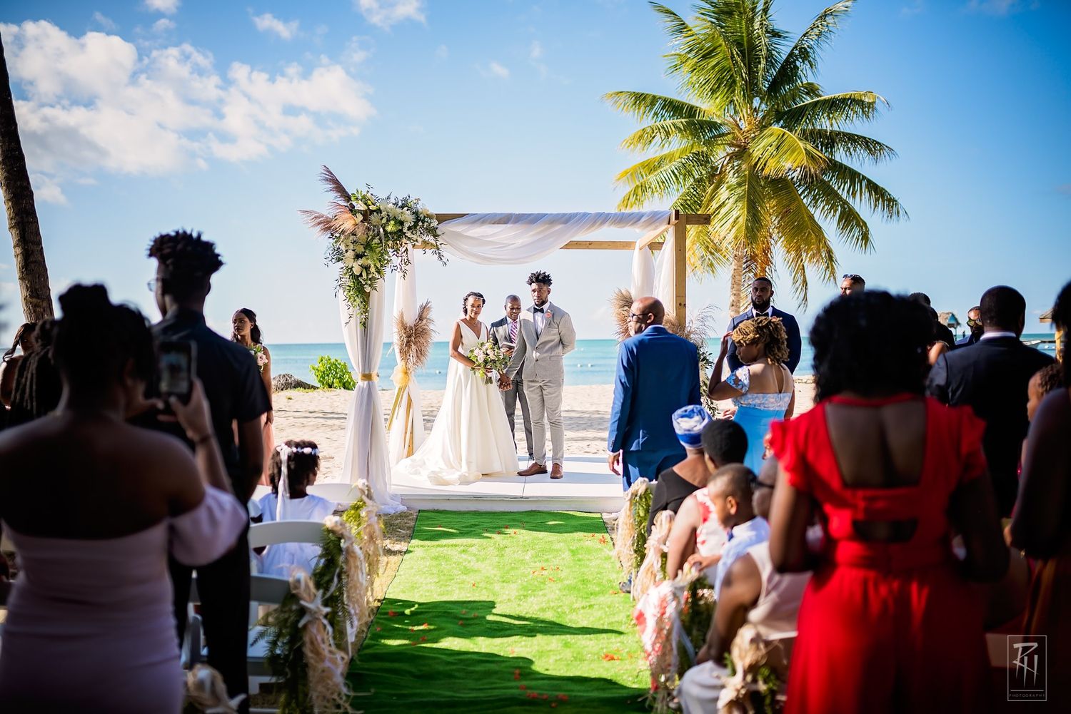 Tropical beach wedding ceremony under palm trees with white arch and guests on green lawn.