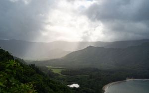 Dramatic sunbeams pierce through storm clouds over a mountainous coastal landscape.