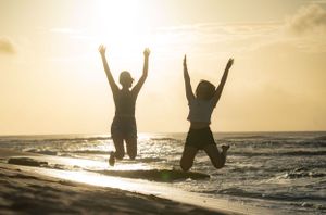 Two silhouettes joyfully jumping on a beach at sunset with ocean waves in the background.