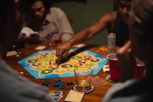 Friends gather around a wooden table playing Settlers of Catan board game with drinks.